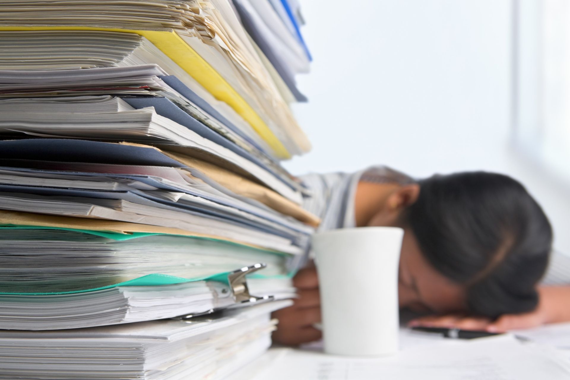Buisness man alseep on his desk which has piled up work on it.