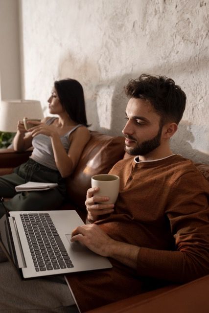 A man with medium skin tone, a beard, and short dark brown hair sits on a sofa next to a woman with medium skin tone and long dark brown hair. The man is holding a coffee cup and looking at a laptop. 