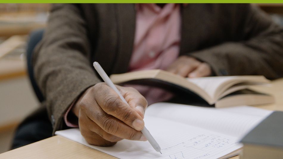 A man’s hand with dark skin tone holds a pen and writes in a notebook in the foreground of the image. In the background his other hand holds the Culturally Responsive School Leadership book open. 