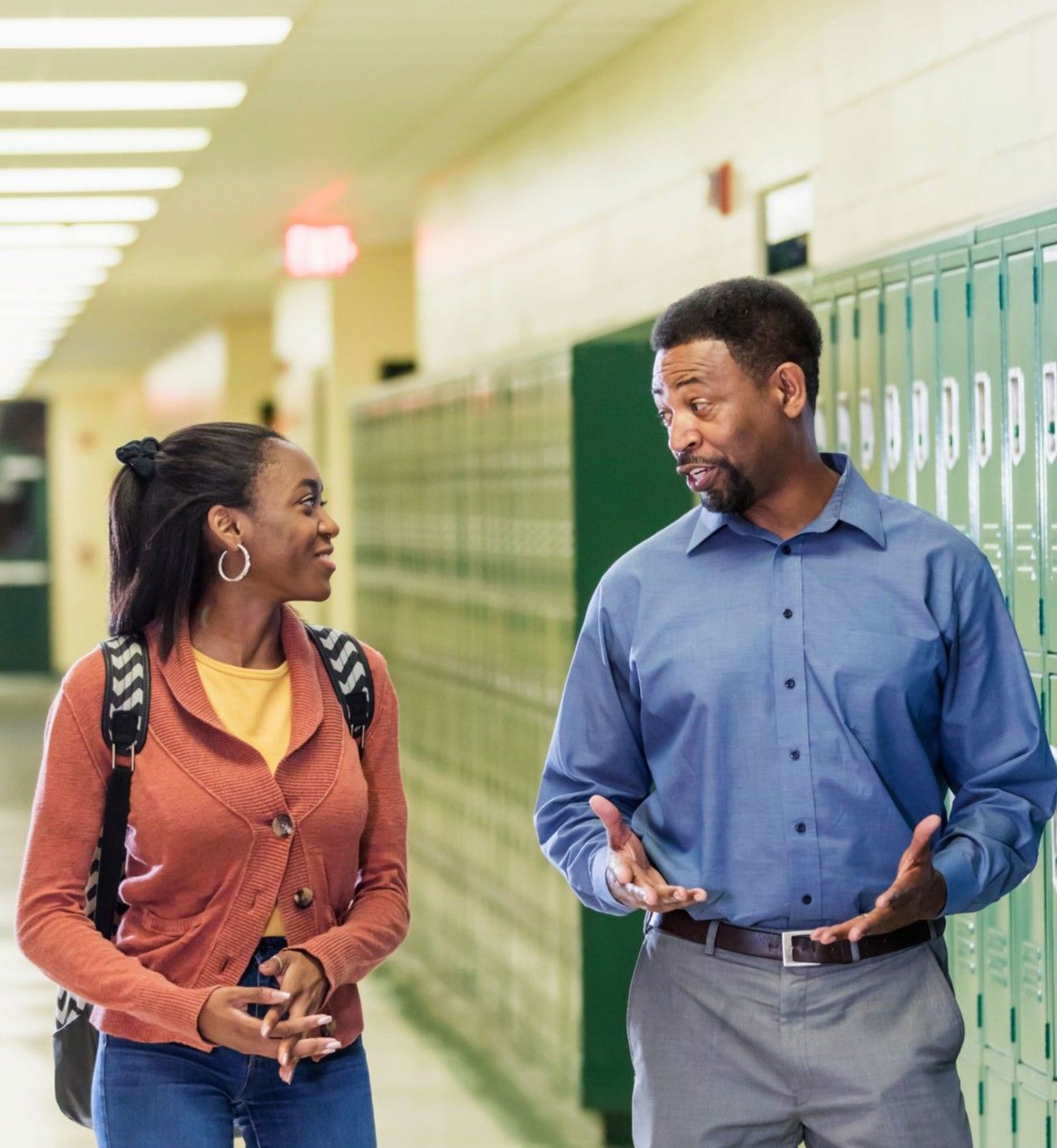 A teacher with dark skin tone stands next to a teenage boy with dark skin tone and headphones around his neck. The teacher is looking at a girl who is in profile. The girl has medium dark skin tone and is sitting with a laptop computer in front of her.  The teacher is wearing a yellow dress and stands with her arm on the boy’s shoulder; both the boy and the teacher smile. 