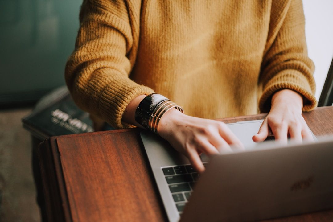 Person typing on a laptop at a wooden desk with a book partially visible in the background.