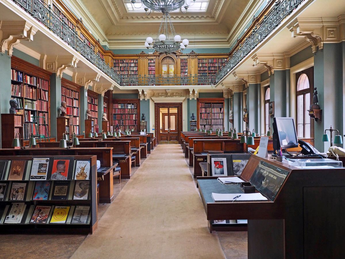 A large historic library reading room with high ceilings, tall bookshelves, long wooden tables with green lamps, and a central aisle leading to double doors.