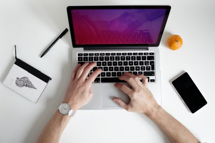 Person using a laptop at a clean desk setup with a notebook, smartphone, pen, and orange placed nearby.