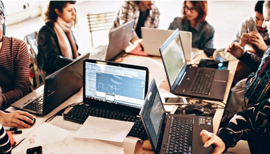 A group of people sitting around a table using multiple laptops and digital devices during a collaborative work or study session.