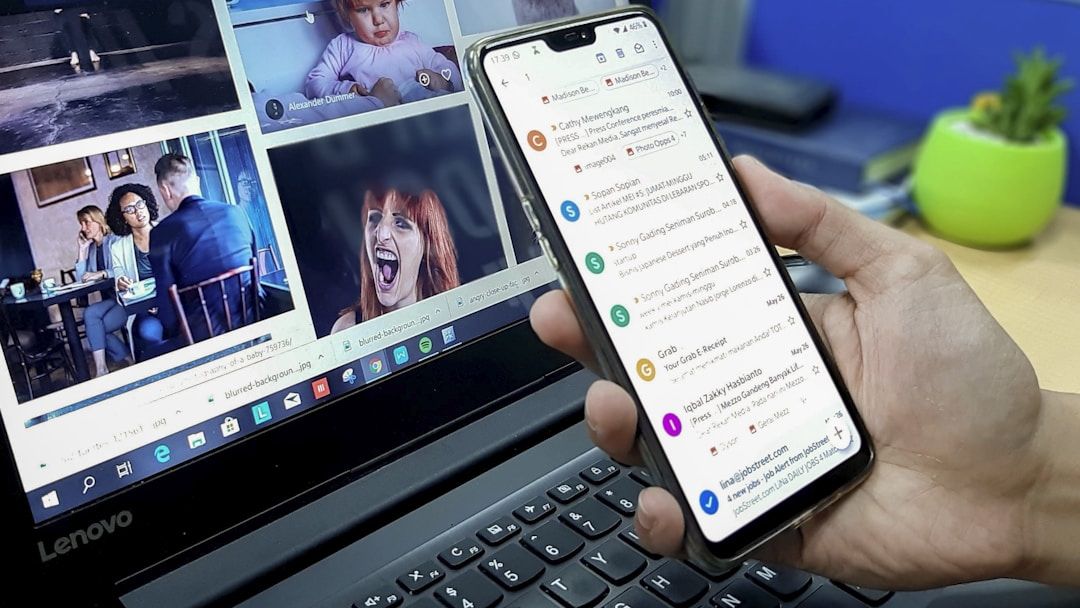 A person holding a smartphone displaying an email inbox while sitting at a laptop that shows several thumbnail images on the screen.