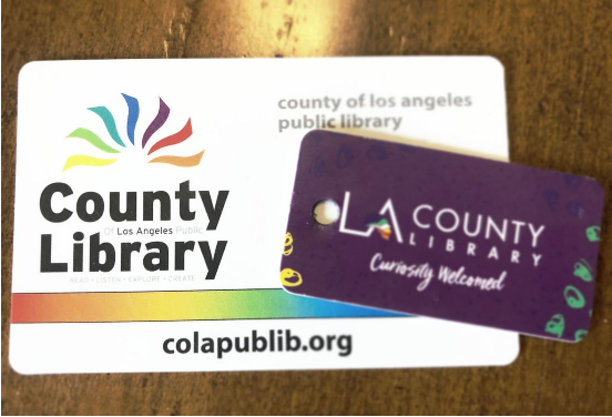 LA County Library cards displayed on a wooden surface, including a large white card with a rainbow design and a smaller purple keychain card.