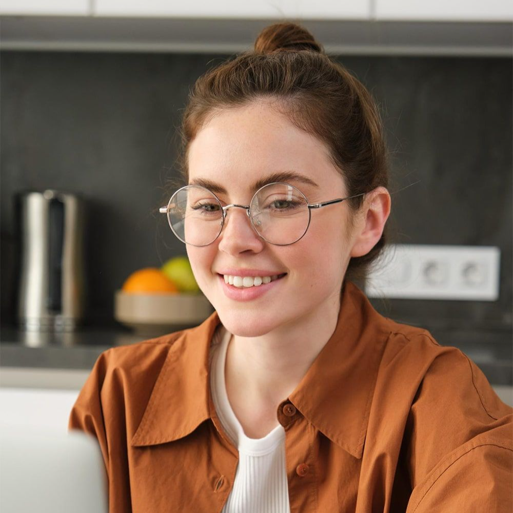 Young woman with glasses on and her hair in a bun smiling while working on her laptop.