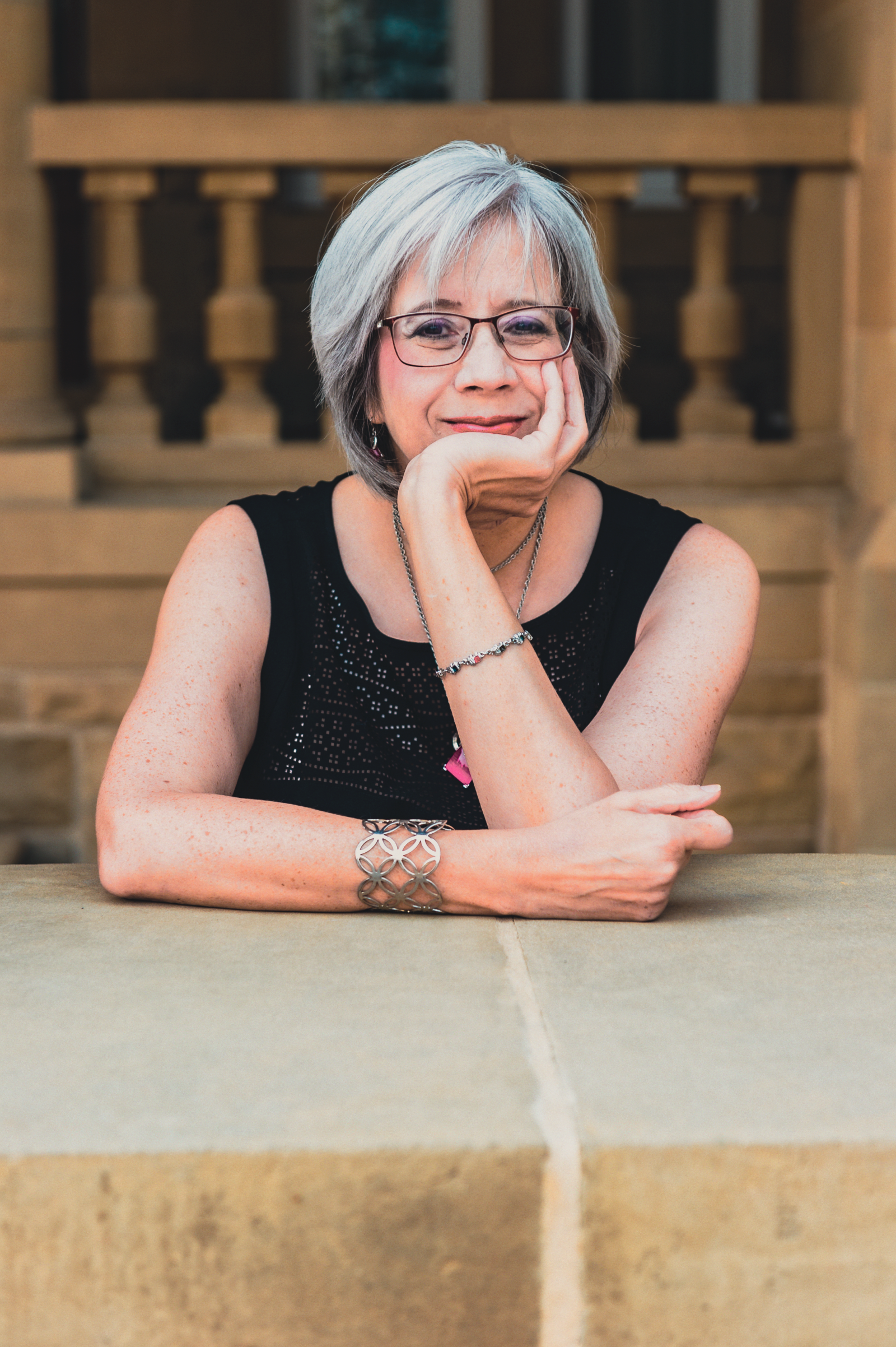 A photo of Rhonda Kronyk, a middle-aged women with silver-grey hair. She is wearing red glasses, a sleeveless black dress, and a chain with a bright pink pendant. She is leaning forward on a short wall and has her chin resting in one hand.