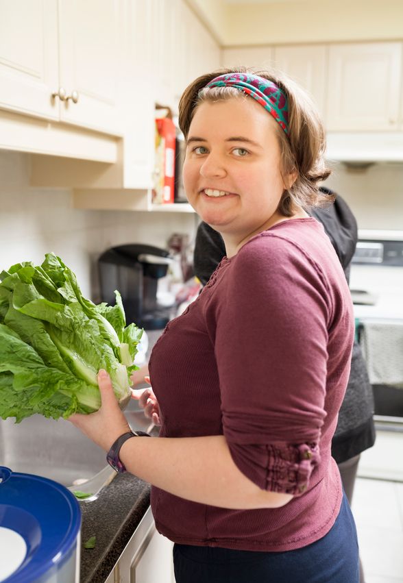 An image of a person facing the camera, wearing a dark red top, standing in a kitchen near a sink. They hold a head of lettuce.