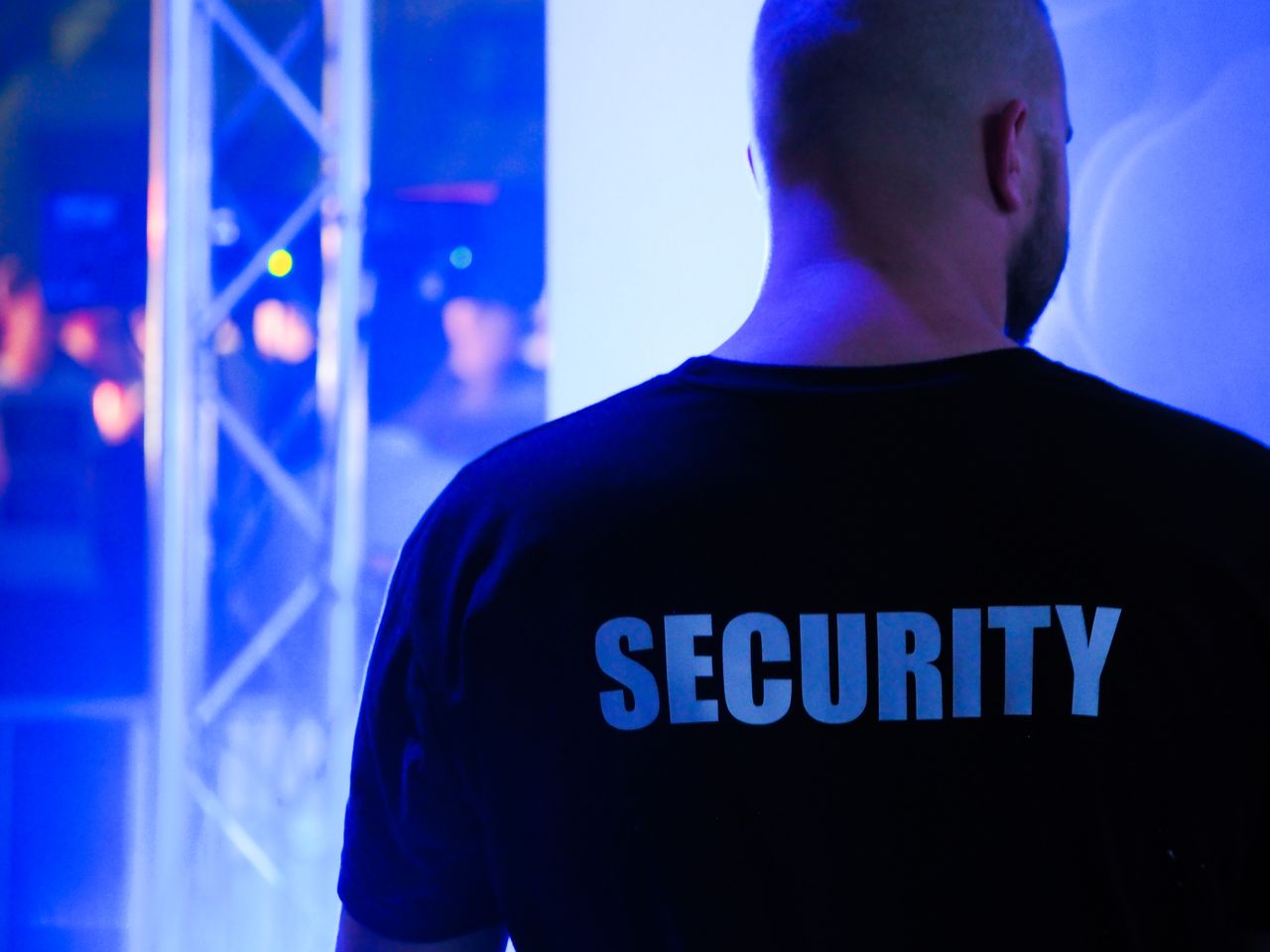 Security guard seen from behind in a black SECURITY shirt at a blue-lit venue