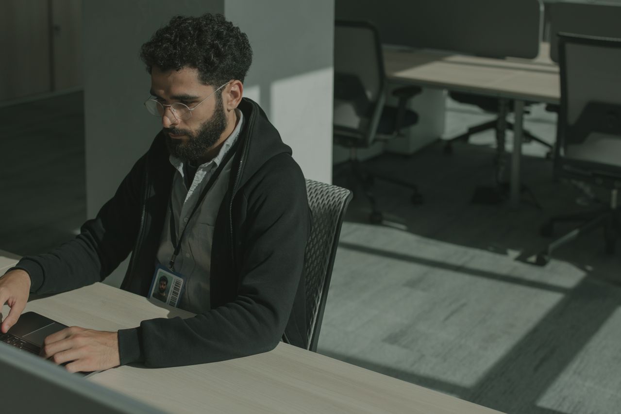 Bearded man in glasses and a dark hoodie with an ID badge typing on a laptop at an office desk