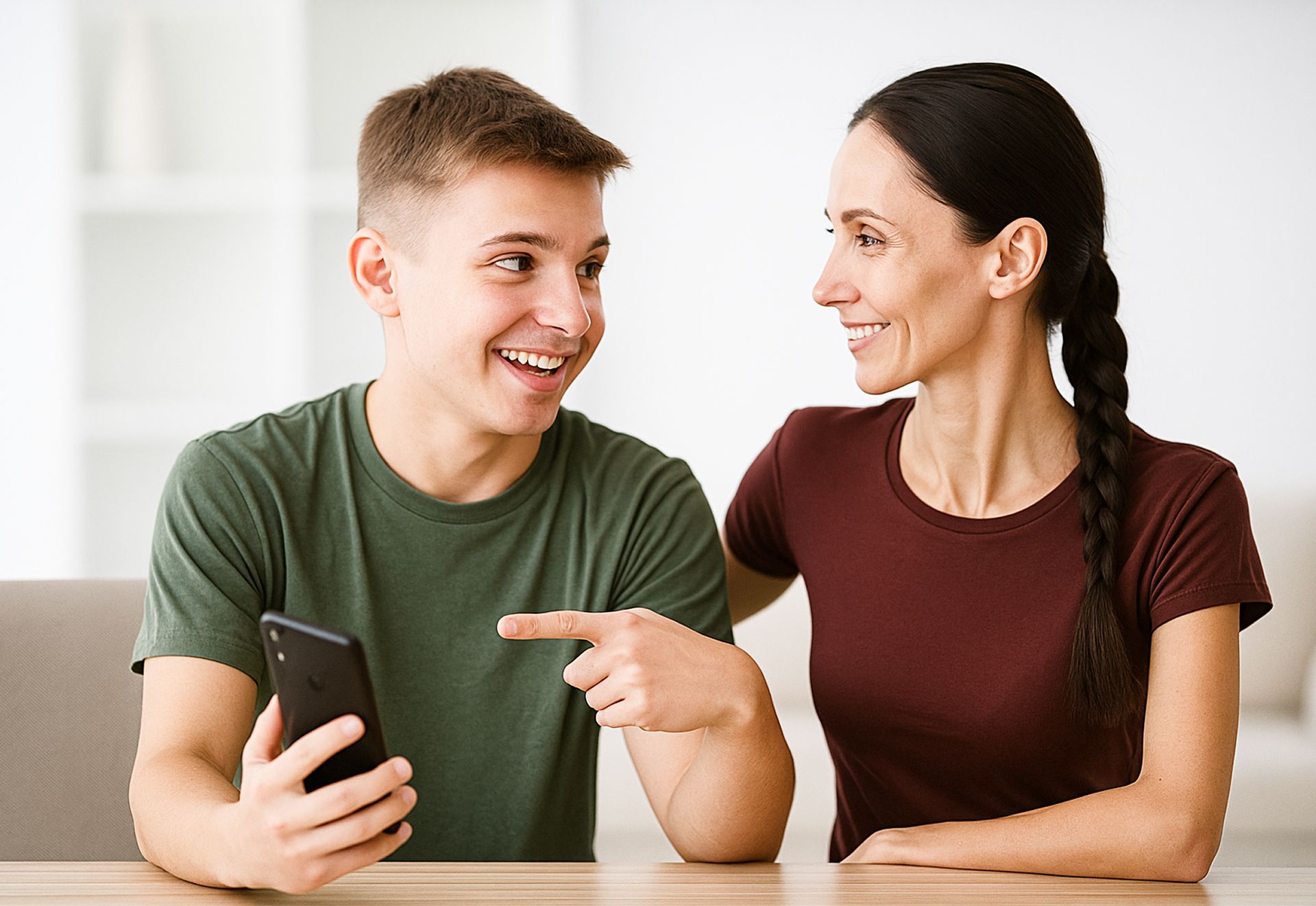Joven sonriente mostrando su teléfono móvil a una mujer que lo observa con interés, ambos sentados frente a una mesa.