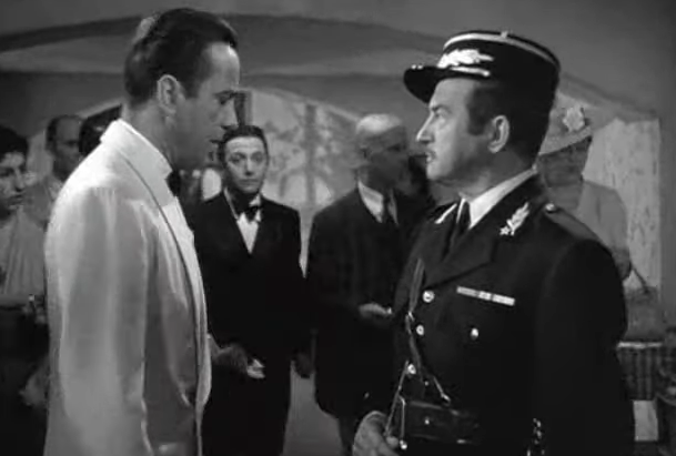 In a black-and-white scene, a man in a white tuxedo faces a uniformed officer in a tense interaction. Onlookers observe in the dimly lit room.