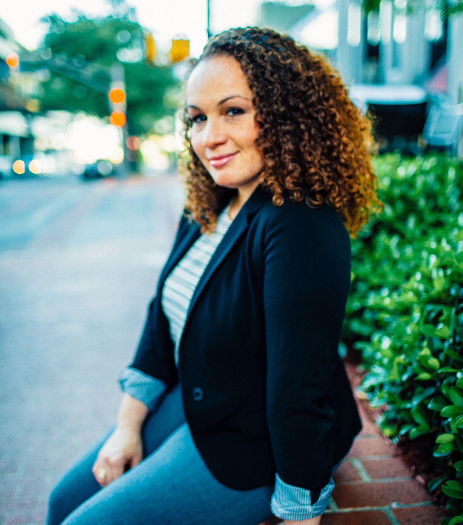 woman with curly hair sitting on brick wall with a closed smile