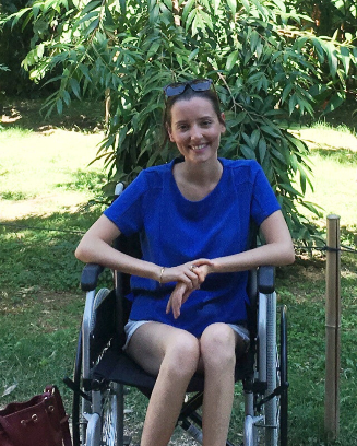 Julie Morin sitting in a wheelchair outdoors, wearing a bright blue shirt and light shorts, with sunglasses resting on her head. She is seated in a garden with green trees and grass in the background.