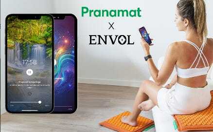 A lady in white workout clothes, sitting on a Pranamat and holding a telephone with the Envol app on the screen.