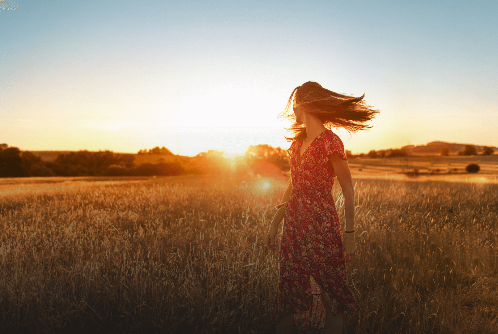 Smiling woman with arms outstretched, in a field with trees on both sides at sunset, glowing in warm golden light.