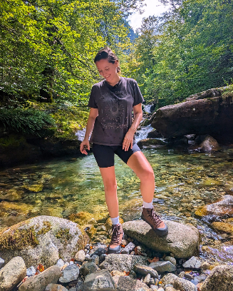 Julie Morin stands on rocks by a clear forest stream, smiling with her head slightly bowed. She is wearing hiking boots, black shorts, and a dark t-shirt, surrounded by lush green trees and sunlight filtering through the leaves.