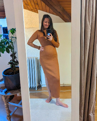 Julie Morin taking a mirror selfie indoors, wearing a long fitted brown ribbed dress with long sleeves. She stands barefoot on a rug, smiling slightly with one hand on her hip, in a cozy room with wooden ceiling beams, with a potted plant in the background.
