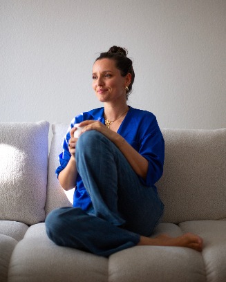 Julie Morin sitting comfortably on a light-colored sofa, holding a white cup in her hands. She is dressed in a bright blue blouse and blue jeans, with her hair tied up in a bun, looking content as warm sunlight falls across her face.