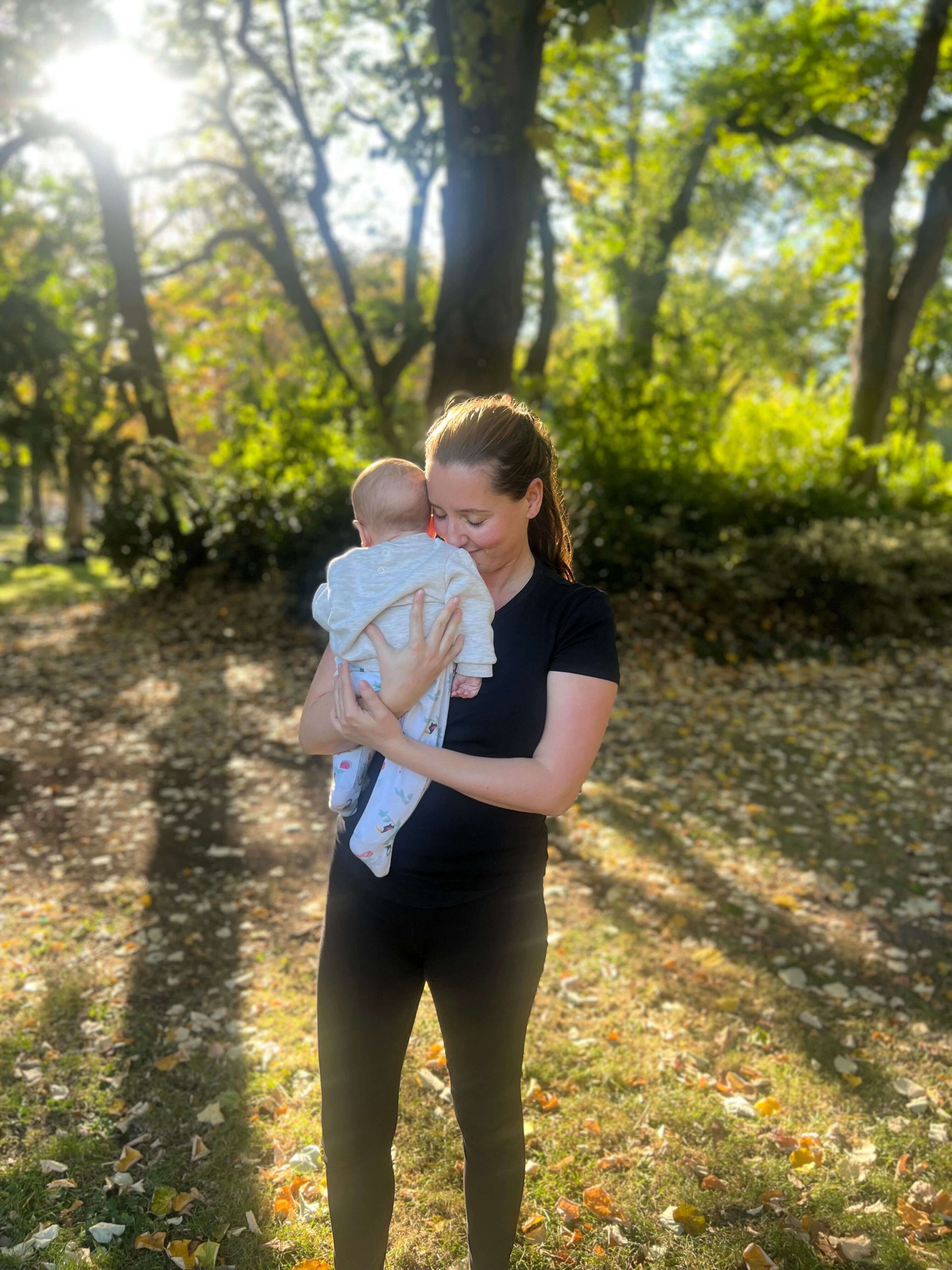 Julie Morin holding a baby close to her chest in a sunlit park, surrounded by trees and fallen autumn leaves, sharing a calm and tender moment.