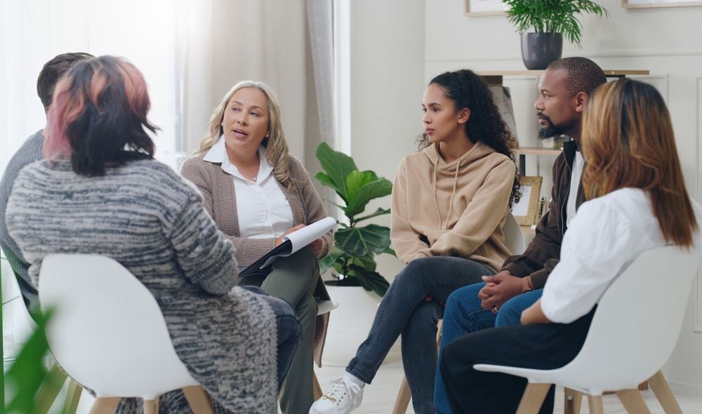 Small group meeting with diverse individuals seated in a cozy, well-lit room. The facilitator leads a discussion, promoting active listening and inclusive dialogue. Ideal for illustrating collaboration, leadership, and personal growth in team settings.