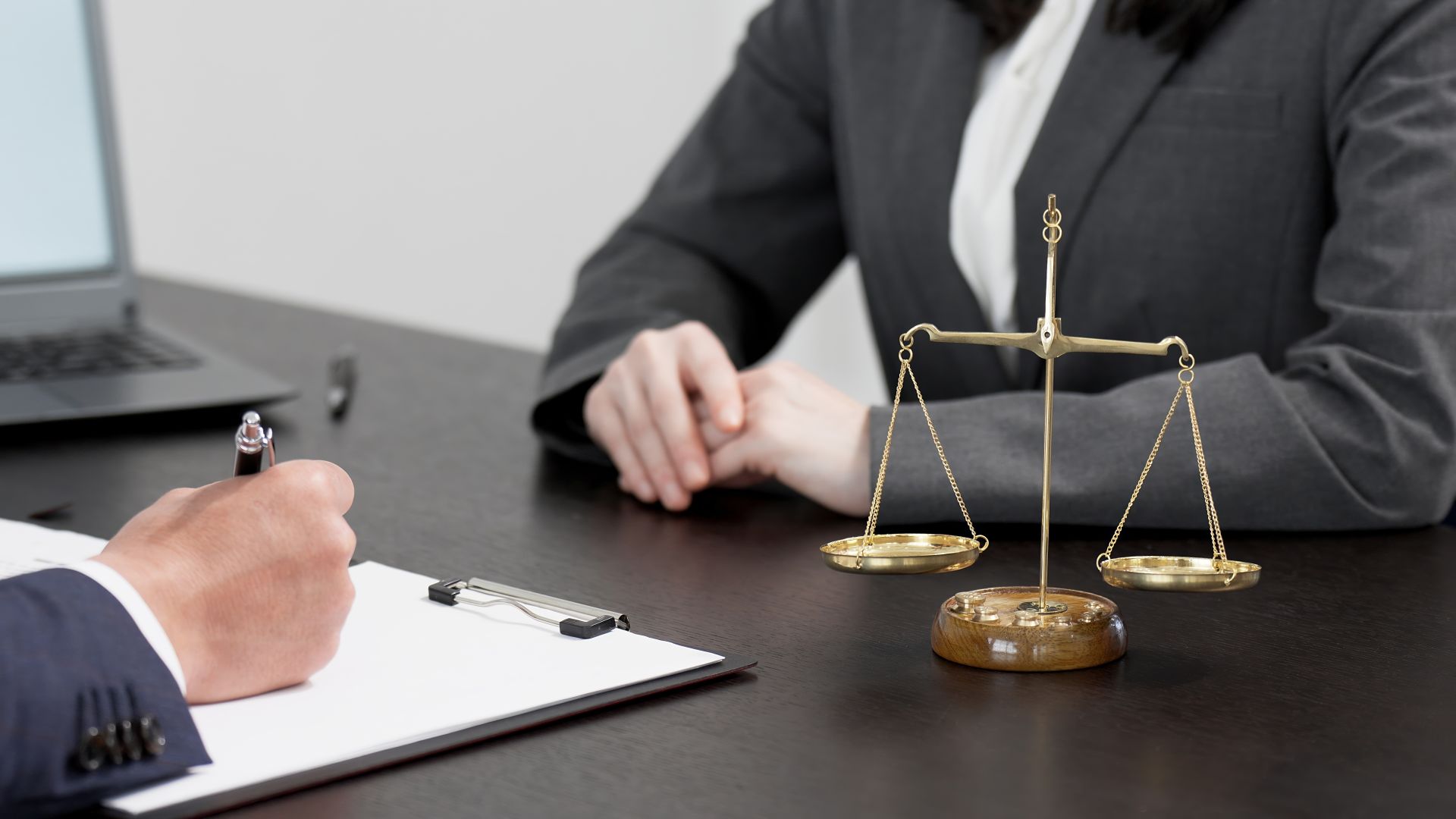 Close-up of a legal professional's desk with a justice scale in the foreground, a clipboard and pen in use, and two individuals in professional attire discussing legal matters.