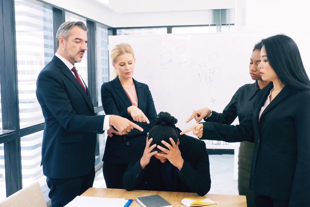 Workplace conflict scene showing a stressed employee seated with their head in their hands while colleagues point fingers in blame. This image highlights toxic work environments, the impact of workplace bullying, and the need for conflict resolution and organizational support.