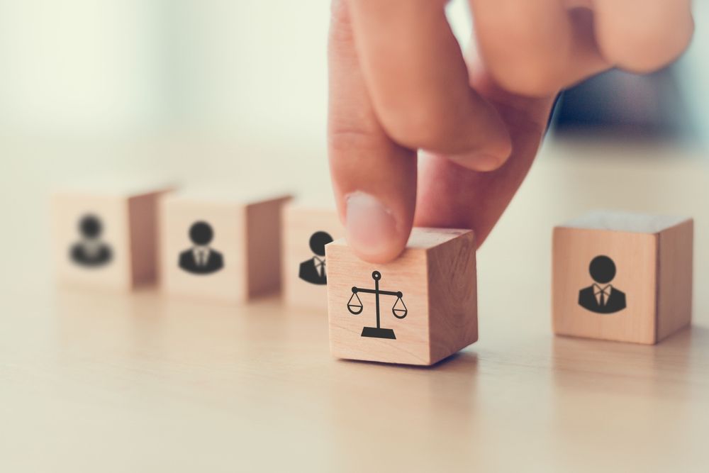 Close-up of wooden blocks on a desk, with a hand selecting a block featuring scales of justice, surrounded by blocks with icons of businesspeople. This image symbolizes fairness, equity, and decision-making in workplace leadership and organizational practices.