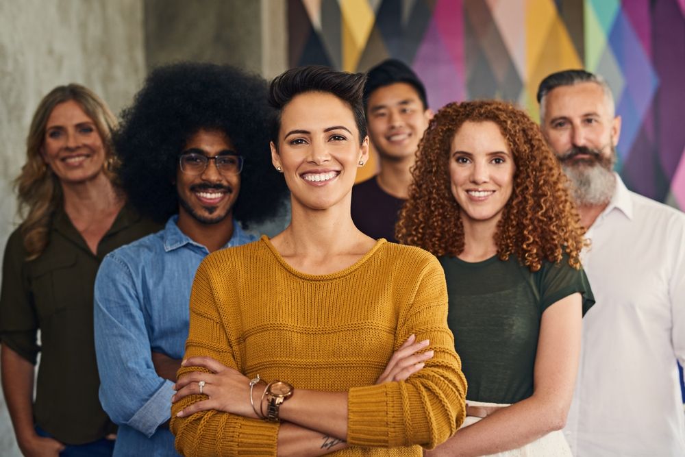 Smiling diverse group of professionals standing together against a colorful geometric background. This image reflects workplace diversity, equity, and inclusion, showcasing collaboration, unity, and representation in professional environments.