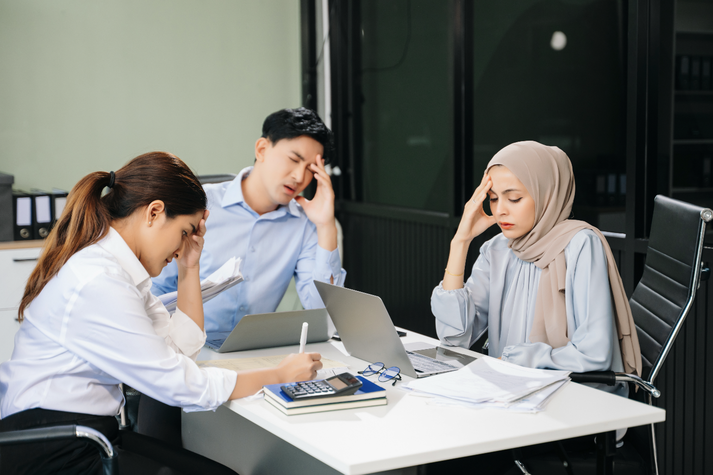 Three coworkers in an office setting looking stressed, with one woman wearing a hijab, all seated around a table covered with documents and laptops.