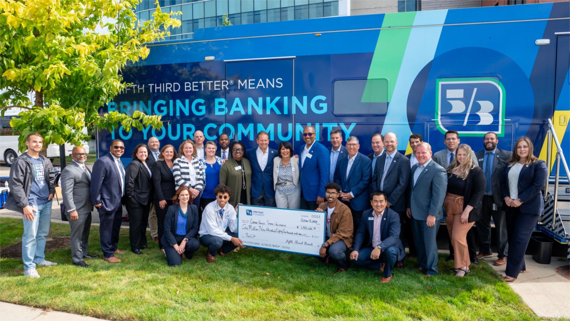 A photo of a big smiling group of people standing in front of a Fifth Third bus. At the front of the photo are two students holding an enormous check from Fifth Third to create the FinLit program.