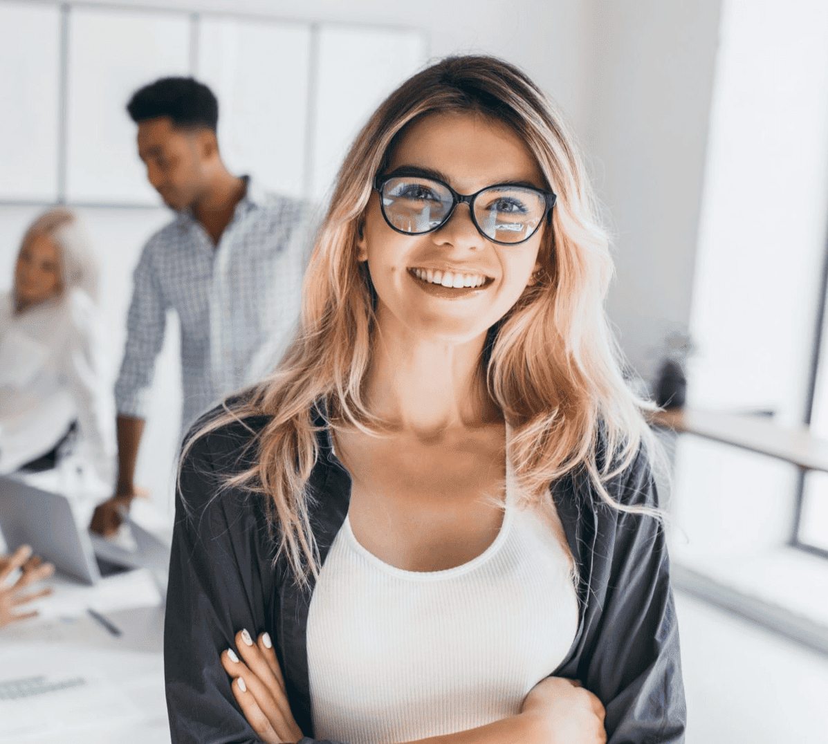 A confident young professional with glasses smiling in a bright modern office, with colleagues collaborating in the background.