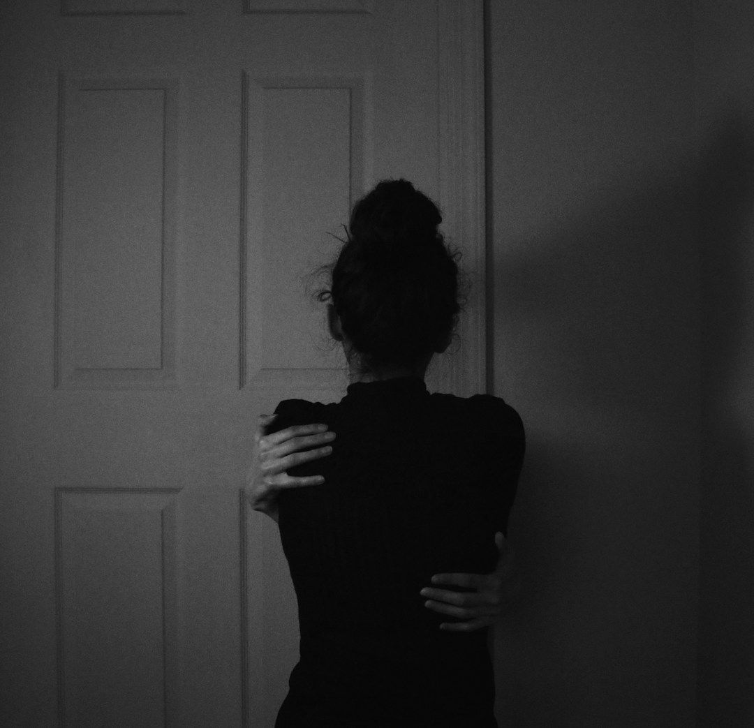 black and white photo of woman in a black sweater standing with her back to us wrapping arms around herself like a hug