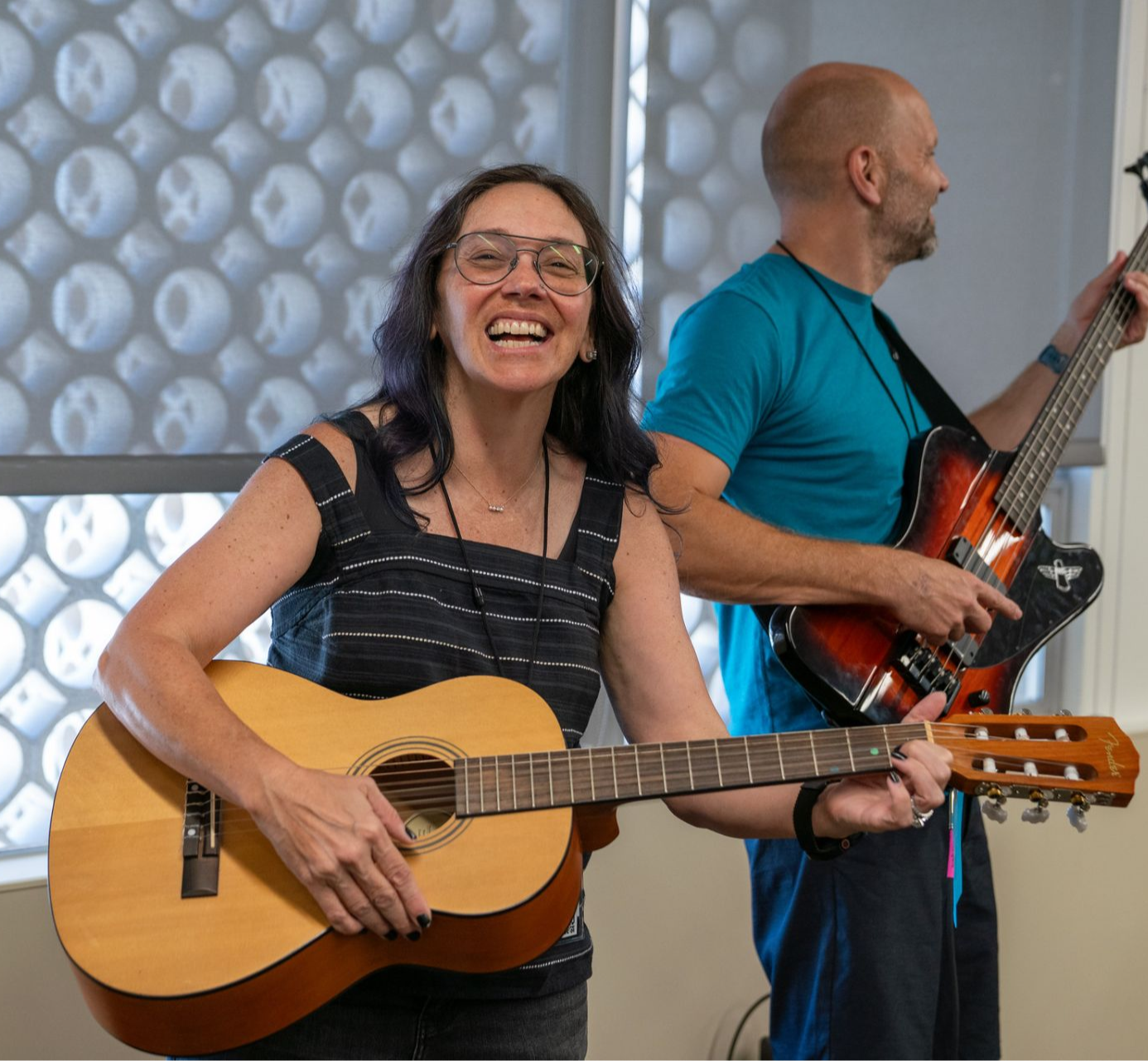 Woman music educator with glasses and brown hair smiling wide at the camera. She is holding an acoustic guitar across her body while standing in front of another music educator who is playing an electric bass guitar while looking away from the camera. 