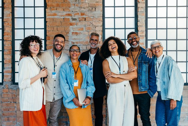 A diverse group of seven smiling professionals are posing for a photo in front of a brick wall with large, grid-patterned windows. They are all wearing lanyards with badges, suggesting a work event or conference. The group includes individuals with varying ages, ethnicities, and styles, creating a sense of inclusivity and collaboration, which might be a focus promoted by a Chief Diversity Officer.