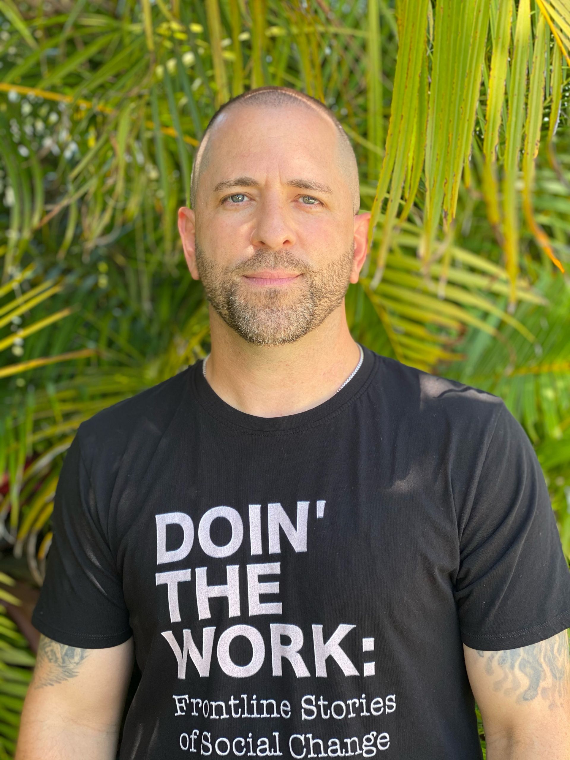 Shimon Cohen, LCSW—Founder and CEO of Doin' The Work LLC, host of the Doin' The Work podcast, therapist, and advocate for justice-based social work—standing in front of greenery wearing a 'Doin' The Work: Frontline Stories of Social Change' t-shirt.
