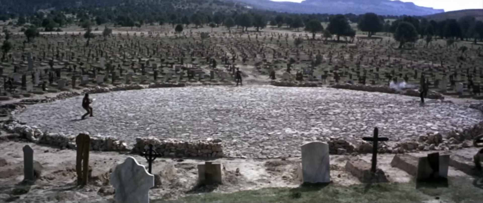 Three figures spread around a wide circular stone clearing in a cemetery, facing off.