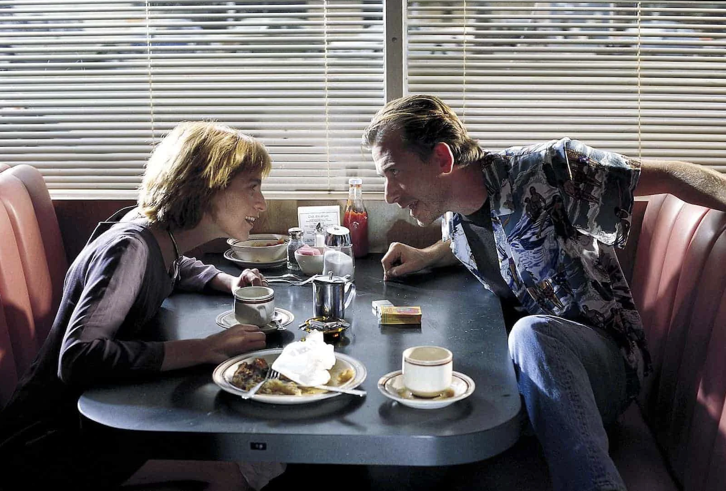 Two people leaning in close across a diner table, faces almost meeting.
