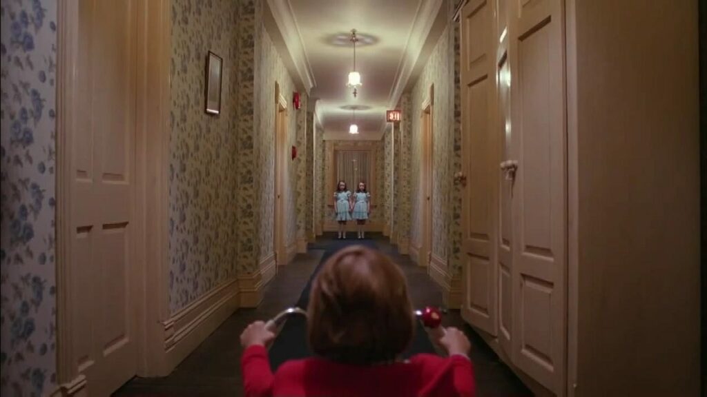 A child on a tricycle at the near end of a long hotel corridor, with two figures standing far away at the other end.