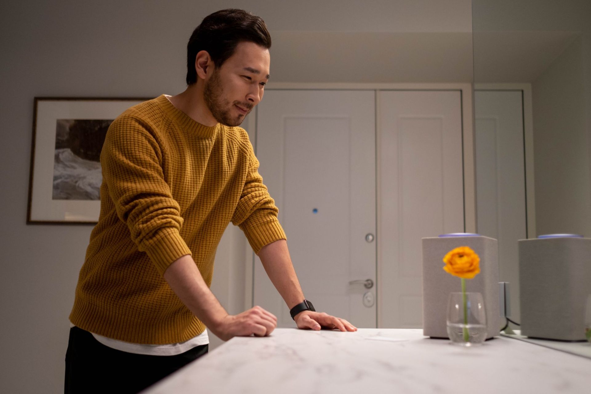 A man at home leaning on a kitchen counter, attentively watching a smart speaker as its light ring glows, relying on an AI product in the moment it responds.