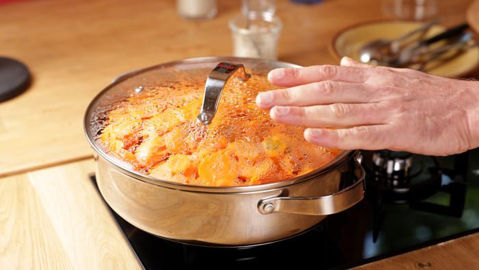 Main posée sur le couvercle d'une casserole de légumes en cuisson à l'étouffée, sur une plaque de cuisson allumée.