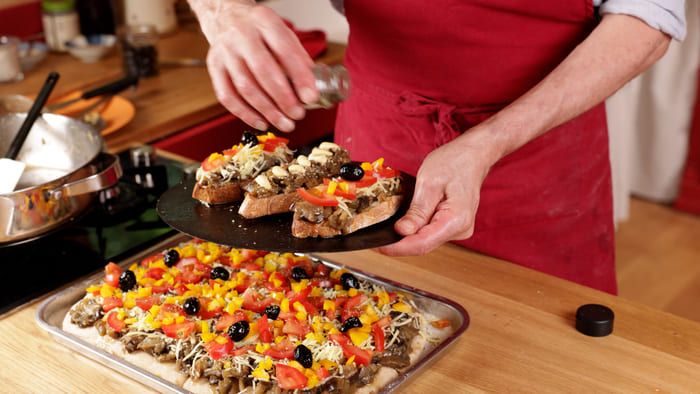 Bruschettes et d'une pizza à pâte levée faite maison, toutes deux garnies de légumes cuits en suée couverte et de légumes frais, sur une table de cuisine.