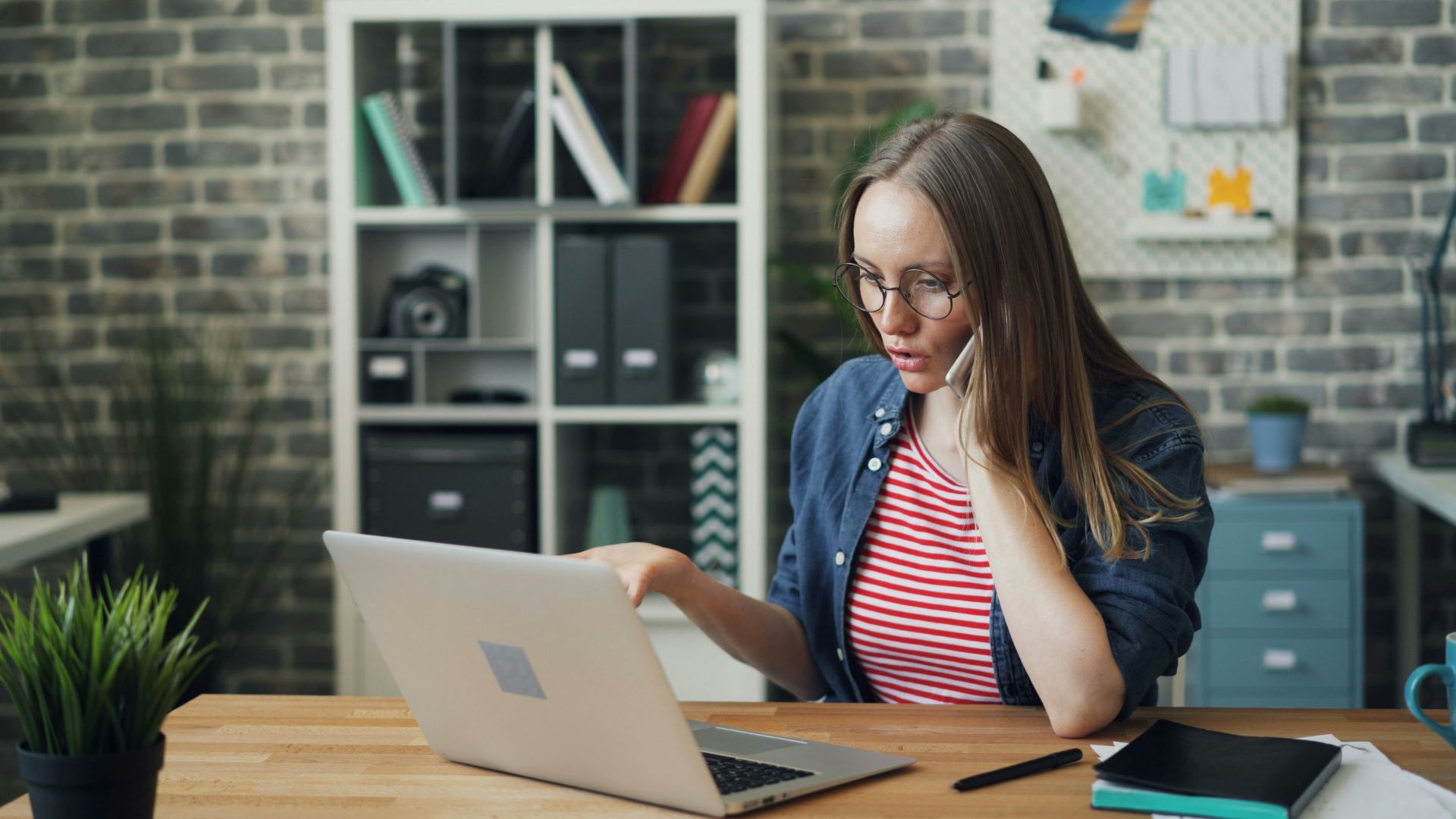 A woman in a casual denim jacket and striped shirt sits at a wooden desk, focused on her laptop