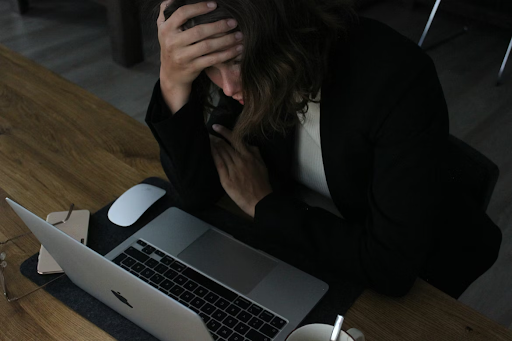 a woman, looking stressed, sits at a wooden desk with a laptop. Their hand rests on their forehead, indicating frustration