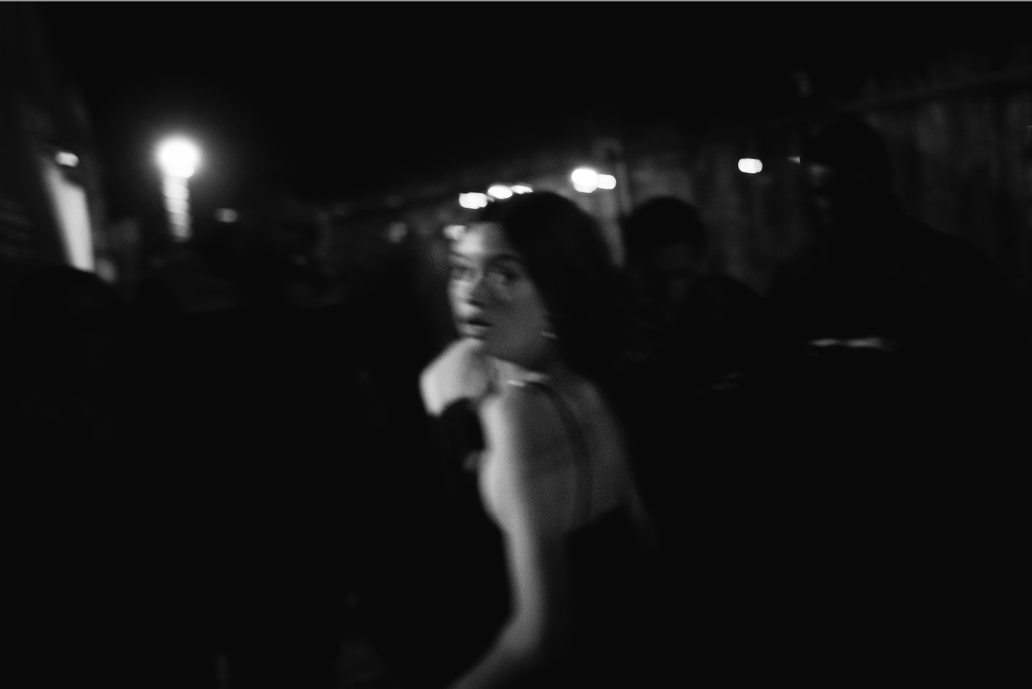  blurred black and white image of a woman in a black dress, looking back over her shoulder at night showing unease