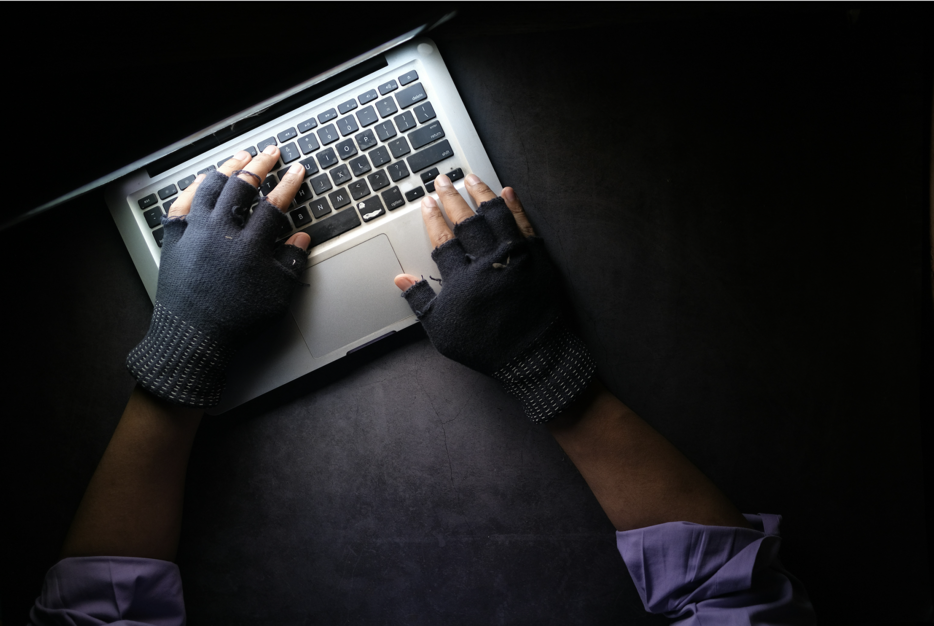 Hands wearing fingerless gloves type on a laptop keyboard in a dimly lit room