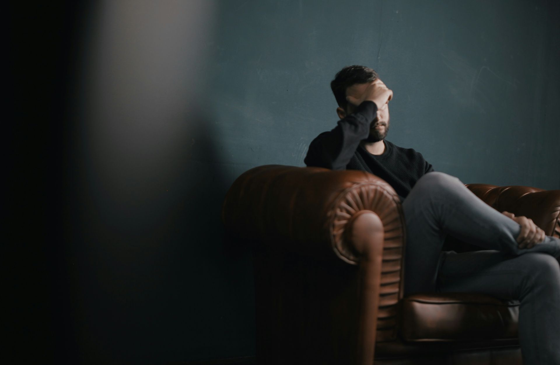  a person sits alone on a brown leather couch, covering their face with one hand looking stressed