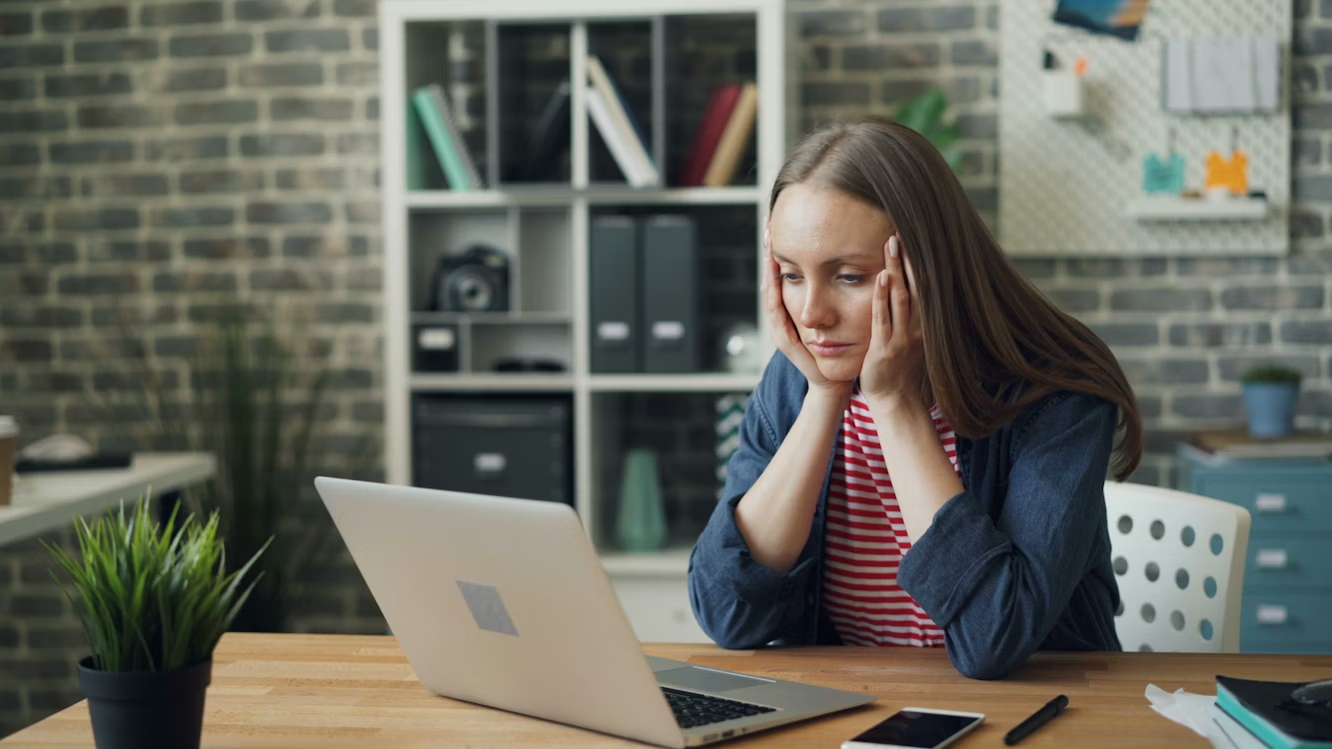 A woman sits at a wooden desk, hands on her temples, looking stressed at a laptop.
