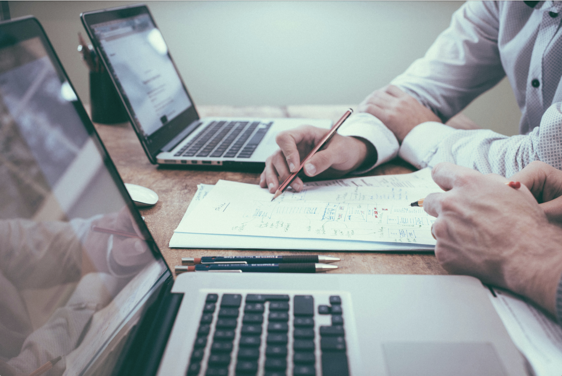 Two people reviewing documents on a wooden table with two open laptops. One points at a paper with a pencil.